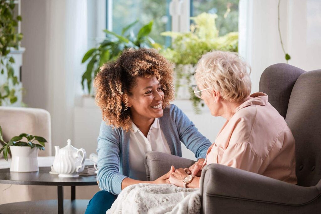 Caregiver warmly chatting with an elderly woman seated comfortably in a bright living space.