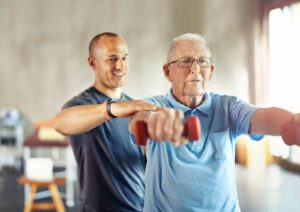 Older man lifting small hand weights assisted by a young male trainer during a fitness session.