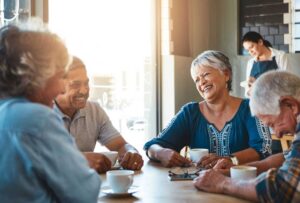 Group of seniors sitting around a table laughing and playing cards near a window.