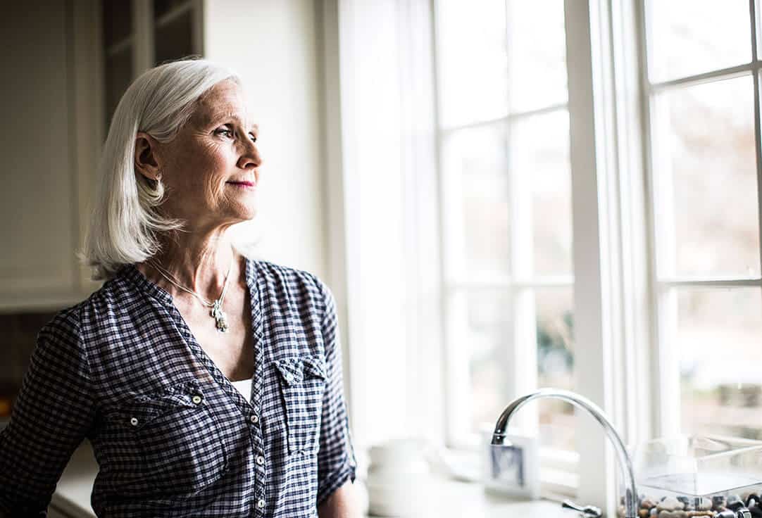 Senior woman standing by her kitchen sink looking out a window deep in tought.