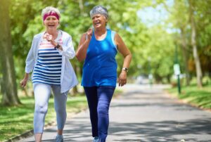 Two senior women laughing and power walking together in a tree-lined park on a sunny day, wearing athletic clothing and headbands